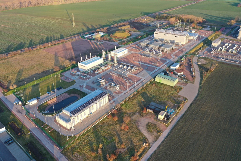 Das Foto zeigt aus einer schrägen Vogel-Perspektive die Gesamtübersicht über die Obertageanlagen eines Untergrundgasspeichers. Im Vordergrund ist eine große Halle zu sehen. Weiter befindet sich im linken Vordergrund ein Feuerwehrlöschteich. Im Mittelteil sind viele Rohrleitungen zu sehen, die teils parallele Leitungsbündel bilden und in Bögen aus und wieder in den Erdboden führen. Weiter sind teils aufrechtstehende Behälter und Industriehallen zu sehen. Im rechten hinteren Teil ist eine große Halle zu sehen an welche sich im Hintergrund elektrische Anlagen (ein Umspannwerk) anschließen, die an eine Hochspannungsleitung angeschlossen sind.