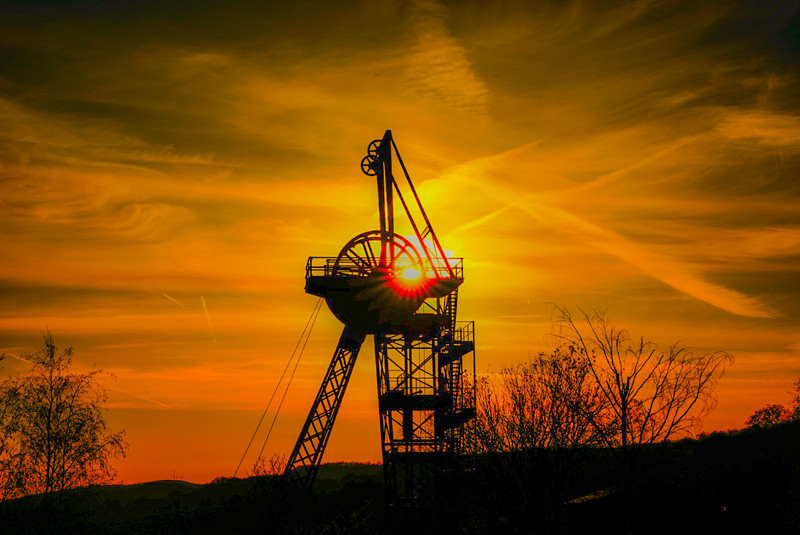 Das Foto zeigt den Förderturm des Röhrigschachtes. Im Hintergrund geht die Sonne unter, so dass sich die Stahlkonstruktion des Förderturms als schwarze Kontur vor dem leuchtend roten Himmel abzeichnet.