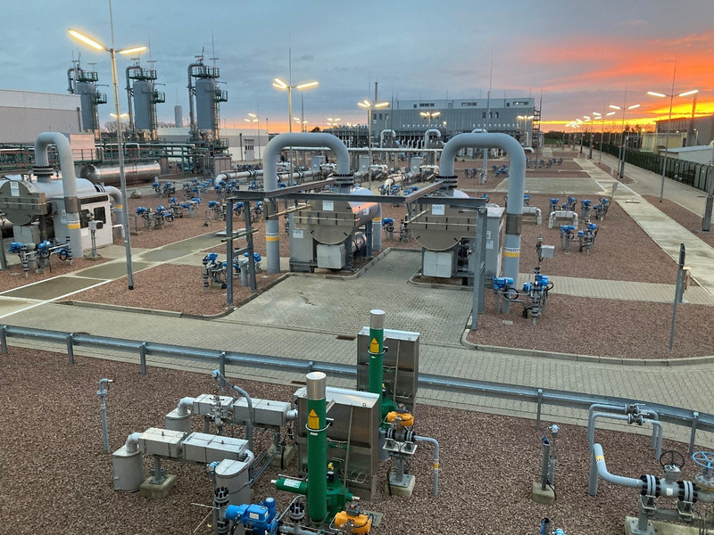 Das Foto zeigt eine Teilansicht der Obertageanlage eines Gasuntergrundspeichers. Der gesamte Platz ist geschottert und dazwischen sind die Fahrwege und Zugangswege gepflastert. Im Vordergrund sind Gasleitungen und Gasarmaturen zu sehen. Dahinter sowie im rechten Bildbereich verlaufen Fahrstraßen und Wege. Dahinter schließen sich liegende, zylindrische Behälter an, in die und aus denen Rohrleitungen führen, die in Bogenform in den Erdboden verlaufen. Im linken Hintergrund sind drei stehende zylindrische Behälter zu sehen. Im linken Bildrand sowie im Hintergrund hinter den Rohrleitungen sind große Industriehallen zu sehen. Ausgeleuchtet wird das Bild durch goldgelb leuchtende Wolken im Hintergrund sowie die eingeschalteten Laternen.